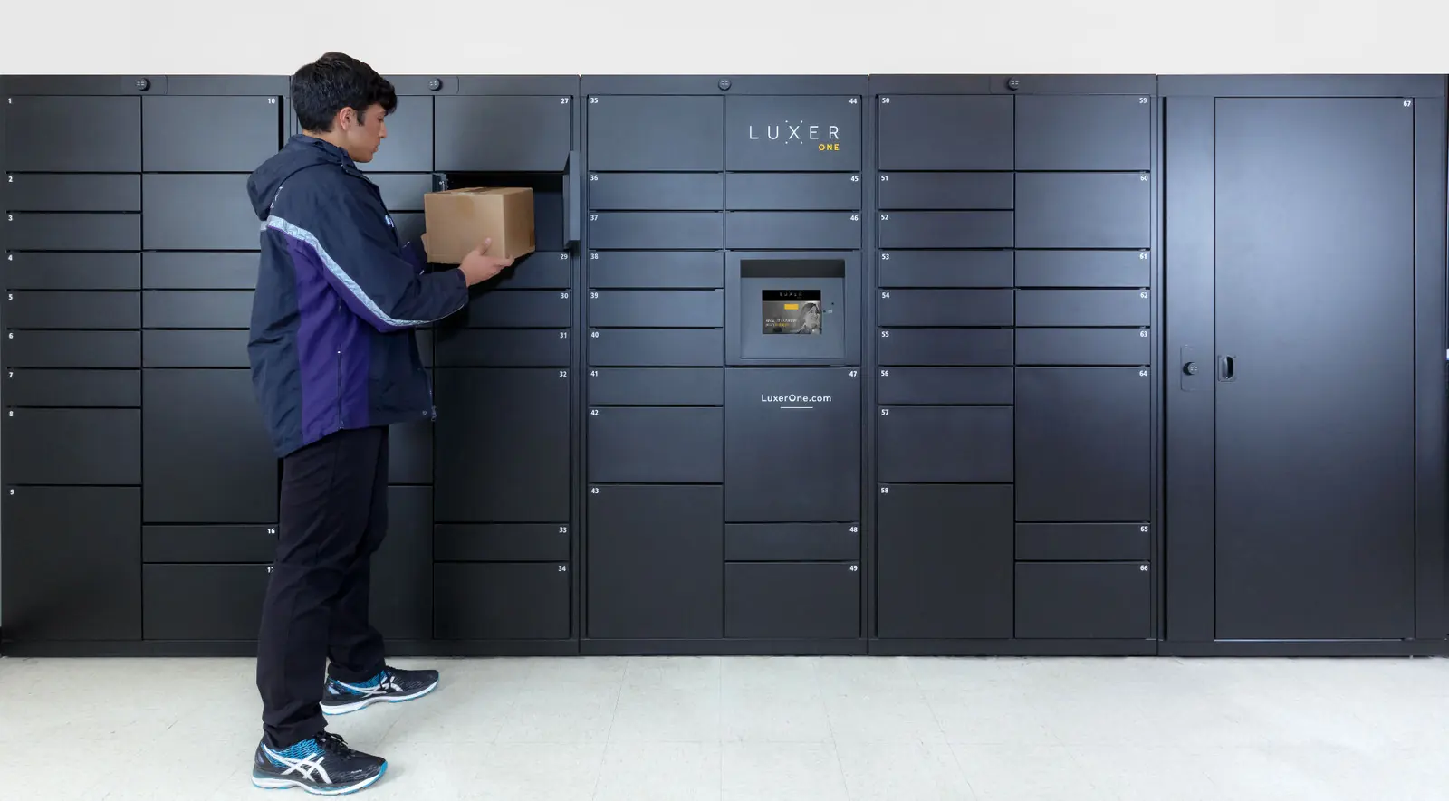 Luxer One indoor electronic package lockers installed in an apartment mailroom — delivery carrier dropping off a resident's package into a smart parcel locker