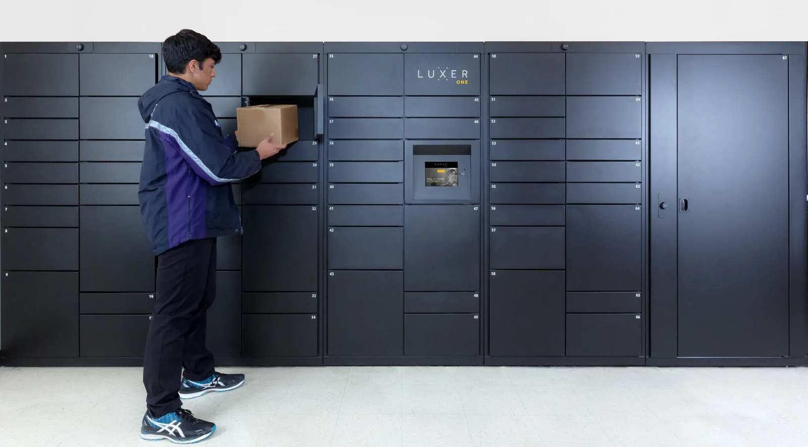 Luxer One indoor electronic package lockers installed in an apartment mailroom — delivery carrier dropping off a resident's package into a smart parcel locker